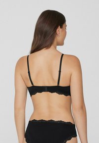 Femme vue de dos portant un soutien-gorge noir à bord en dentelle et un ensemble assorti avec des bretelles réglables et des boucles d'oreilles en argent, sur fond gris.