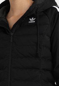 Svart vadderad jacka med dragkedja fram, huva och justerbara snören. Har den vita Adidas-logotypen på bröstet och textil med struktur.