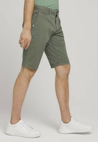 Shorts vert texturés arrivant au genou avec des poches avant et des passants de ceinture. Associés à des baskets blanches, présentés sur un fond uni.
