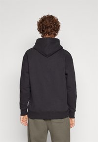 Felpa nera con cappuccio, dal taglio morbido e rilassato, con polsini e orlo a coste. Tessuto soffice con una texture liscia, vista dal retro.