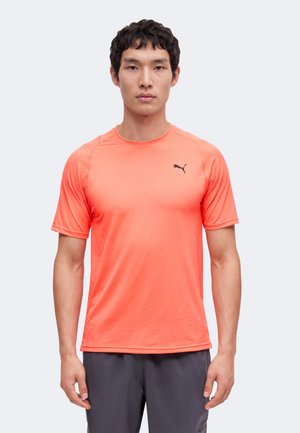 Mand iført en klart orange Puma atletisk t-shirt med korte ærmer og mørkegrå shorts står foran en ensfarvet baggrund.