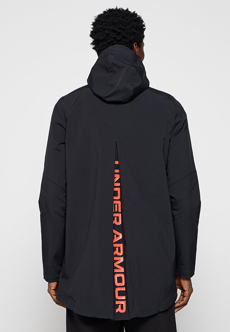Veste noire avec capuche et évent à l'arrière. Comprend un logo « UNDER ARMOUR » rouge le long du dos. Fabriquée en matériau lisse et léger.