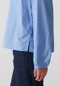 Camicia azzurra a maniche lunghe con taglio dritto, caratterizzata da spacchi laterali, polsini con bottoni e una texture liscia. Indossata sopra jeans scuri.