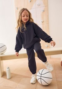 Sweat à capuche noir et jogging assorti, avec des poignets élastiques, portés par un enfant. Le décor comprend un ballon de basket et une bouteille d'eau.