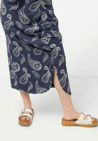 Laivasilta sininen hame, jossa on paisley-kuvio, ja sivuhalkio. Valkoiset sandaalit puupohjalla ja metalliset yksityiskohdat soljessa.