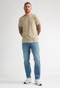 T-shirt beige à manches courtes avec un col rond, associé à un jean en denim bleu clair et des baskets blanches avec des accents foncés.