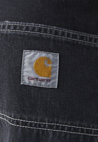 Mørk denimstoff med et kvadratisk grått merke med en gul logo og rød tekst, sydd langs kanten med kontrastvevd hvit tråd.