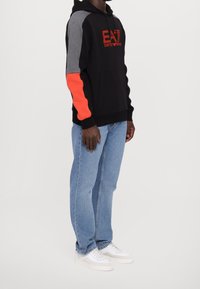 Sudadera negra con mangas grises y naranjas, con el logo bordado "EA7". Combinada con jeans de color azul claro y zapatillas blancas.