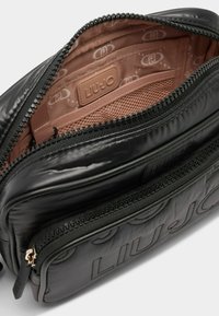 Melnā kvadrātveida soma ar vairākiem ar rāvējslēdzējiem aprīkotiem nodalījumiem, ar gludu ārējo virsmu, mīkstu iekšējo oderi un iegravētu zīmolu.