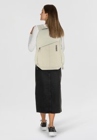 Sac à dos Thule gris clair avec une poche zippée, des bretelles rembourrées et une forme courbée, porté par une personne en jupe en denim noir et des chaussures.