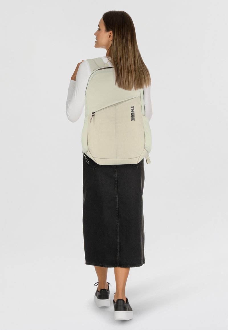 Sac à dos Thule gris clair avec une poche zippée, des bretelles rembourrées et une forme courbée, porté par une personne en jupe en denim noir et des chaussures.
