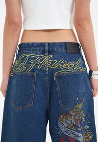 Denim shorts with yellow embroidered text "Ed Hardy" on the back, featuring colorful rhinestone tiger design on the right pocket.