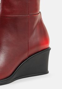 Stivale alla caviglia in pelle rossa liscia con tacco a zeppa nero, cuciture dettagliate e punta leggermente affusolata su sfondo bianco.