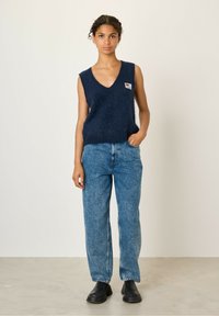 Marineblauwe mouwloze trui in een zachte, fuzzy textuur met een diepe V-hals en een klein kleurrijk patch; gecombineerd met hoge blauwe jeans en zwarte klompen.