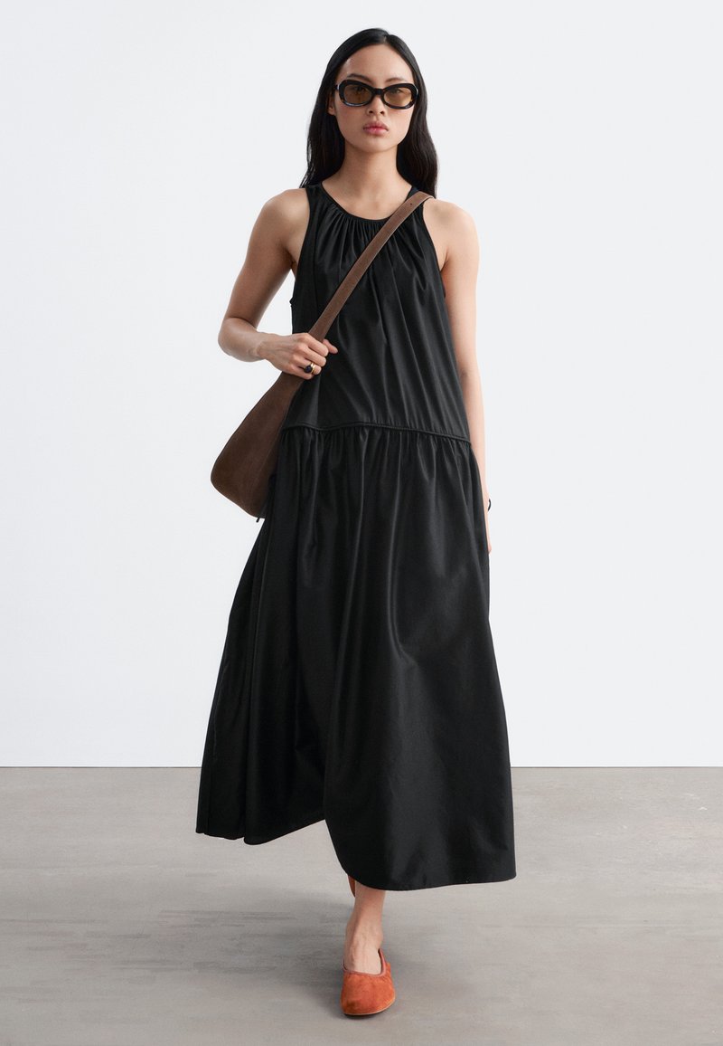 Femme en longue robe noire sans manches, sac bandoulière marron, grandes lunettes de soleil et chaussures orange à enfiler marchant à l'intérieur sur un sol gris.