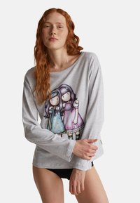 Junge Frau mit langen roten Haaren, die ein graues Langarmshirt trägt, auf dem zwei illustrierte Mädchen in bunten Kleidern abgebildet sind, kombiniert mit schwarzen Hosen.