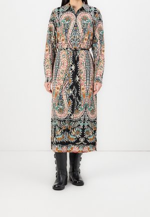 Femme portant une robe midi à manches longues avec un motif floral, associée à des bottines noires à boucle mi-mollet, debout devant un fond blanc.