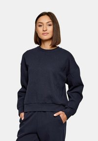 Felpa blu scuro con un design texturizzato caratterizzato da lettere in rilievo. Collo, polsini e orlo a costine. Il modello la indossa con pantaloni abbinati.