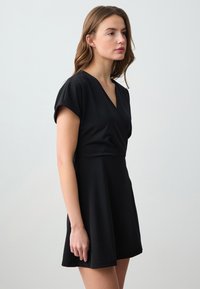 Schwarzes Kleid aus weichem Stoff, mit V-Ausschnitt, kurzen Ärmeln und einem ausgestellten Rock. Das Gesamtdesign ist schlicht und elegant.