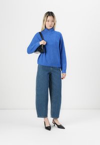 Maglione azzurro lavorato a maglia con colletto alto, abbinato a jeans in denim blu a vita alta e larghi. Il modello tiene una piccola borsa nera e indossa tacchi neri.