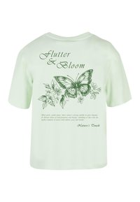 Lys grøn bomuld t-shirt med et sommerfugl- og blomsterdesign på ryggen, med teksten "Flutter & Bloom" i grøn skrift.