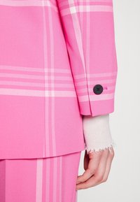 Gros plan sur une main à côté d'une veste de costume à carreaux roses et d'un pantalon assorti, retroussé avec un seul bouton noir sur une manche beige clair effilochée.