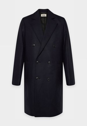Un long manteau noir croisé, avec de larges revers, six boutons et des poches latérales, sur un fond clair.