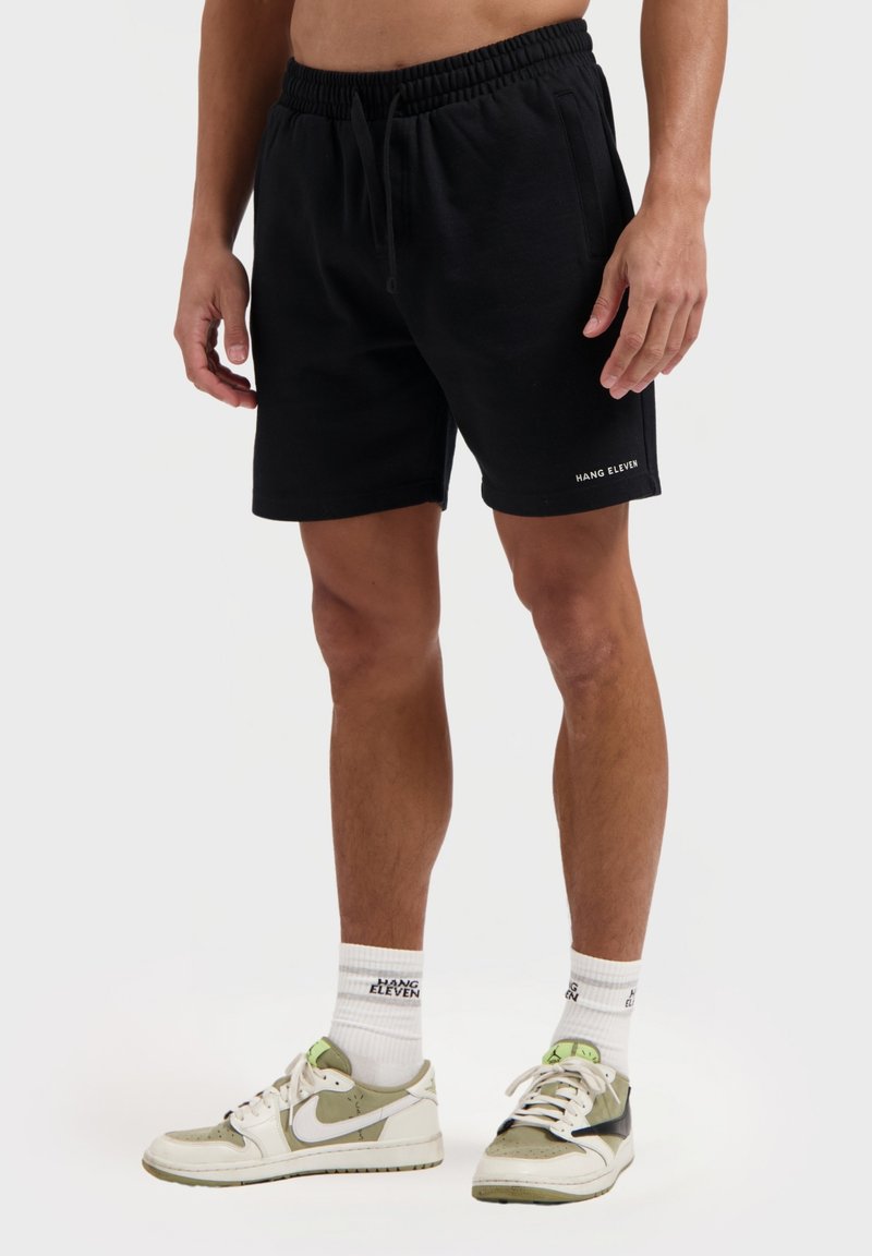 Zwarte katoenen shorts met een elastische tailleband, zijzakken en het "HANG ELEVEN" logo in het wit aan de onderlinkerkant. G gedragen met witte sokken en schoenen.