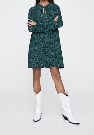 Green floral dress with a round neckline and tie detail. Long sleeves and relaxed fit. Paired with white cowboy boots.