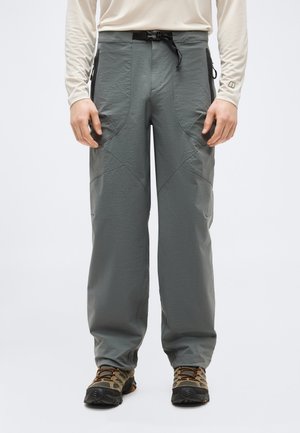 Homme portant un pantalon de randonnée gris clair avec des poches latérales zippées et des chaussures de randonnée, debout devant un fond uni.