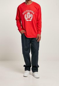 Red long-sleeve t-shirt with white "SOUTHPole" graphic and laurel design, paired with dark blue jeans and white sneakers.