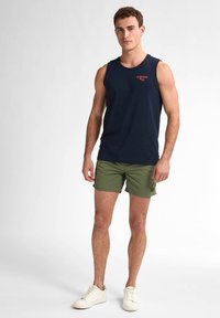 Canotta blu navy con logo rosso, abbinata a pantaloni corti verde oliva. Il modello indossa sneakers bianche, mostrando un outfit semplice e casual.