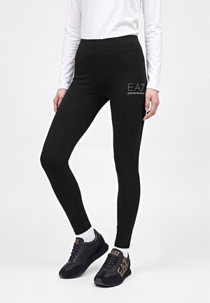 Жена, носеща черни клинове EA7 Emporio Armani и съвпадащи черни маратонки с бели чорапи на едноцветен фон.