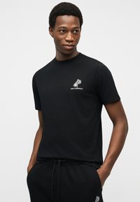 Νέος άνδρας με μαύρο κοντομάνικο μπλουζάκι και παντελόνια με λευκά λογότυπα φοίνικων που ταιριάζουν, με τα χέρια στις τσέπες, κοιτάζοντας δεξιά σε απλό φόντο.