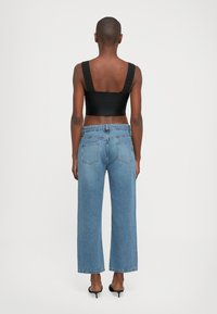 Woman with short hair wearing black crop top, high-waisted loose blue jeans, and black heeled sandals facing away against white background.