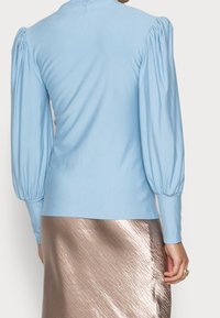 Light blue pullover top with puffed long sleeves, soft texture, fitted silhouette, and crew neckline, paired with a shiny bronze skirt.