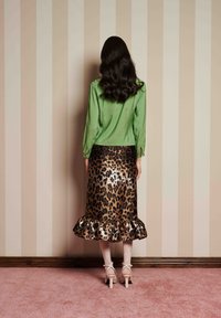 Blusa verde de manga longa, saia metálica de padrão leopardo com bainha em babados e saltos de cor clara, contra um fundo de papel de parede e tapete listrados.