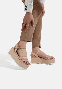 Sandali piattaforma beige con una tracolla testurizzata, cinturino alla caviglia regolabile e suola in gomma bianca. Presentano un dettaglio accentato rosa arricciato.