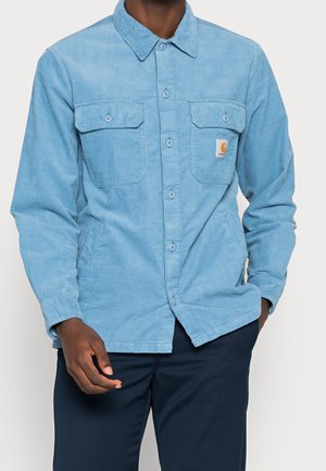 Muž nosící světle modrou kordurovou košili s knoflíky a dvěma náprsními kapsami, tmavě modré kalhoty, ruka v kapse.