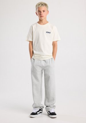 Crème-kleurig t-shirt met korte mouwen met de tekst "VENICE", gecombineerd met lichtgrijze joggingbroek en zwarte sneakers met witte accenten. Eenvoudige, casual outfit.
