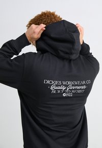 Sudadera negra de tejido suave con capucha ajustable. En la parte trasera tiene texto blanco que dice "Dickies Workwear Co. Prendas de calidad construidas para resistir 1922."