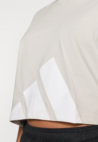 Šviesiai smėlio spalvos trumpomis rankovėmis trumpintas medvilninis marškinėlis su baltais grafiškais dryžiais kairėje pusėje; lygi tekstūra ir laisvas siluetas.