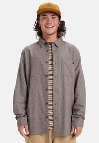 Bruine button-up shirt in een gestructureerde stof met een borstzak, gelaagd over een gestreept T-shirt. Draag met beige broek en een gele pet.
