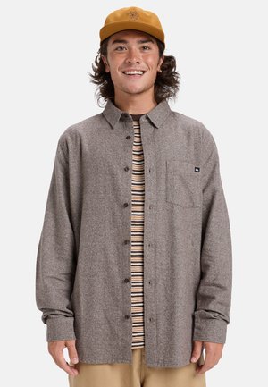 Bruine button-up shirt in een gestructureerde stof met een borstzak, gelaagd over een gestreept T-shirt. Draag met beige broek en een gele pet.