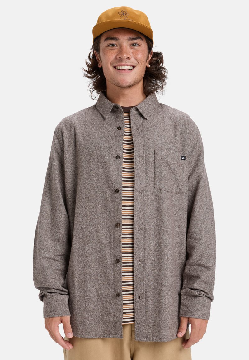Bruine button-up shirt in een gestructureerde stof met een borstzak, gelaagd over een gestreept T-shirt. Draag met beige broek en een gele pet.