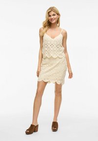 Robe en dentelle crochet beige avec un ourlet en dentelle festonné et un décolleté en V, design superposé, à fines bretelles et silhouette ajustée.