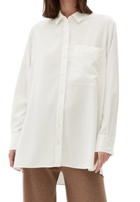 Biała koszula z długim rękawem zapinana na guziki, z kieszonką na piersi, wykonana z gładkiego materiału, z klasycznym kołnierzykiem i luźnym krojem.