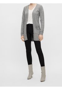 Person wearing a white V-neck top, long gray cardigan, black skinny jeans, and light gray ankle boots, standing on a white background.