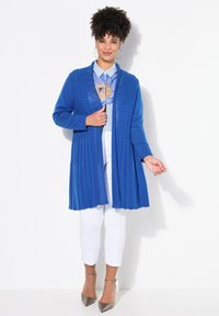 Cárdigan azul real con punto texturizado, de frente abierto y mangas largas. Llevado sobre una camisa azul claro y pantalones blancos, combinado con tacones metálicos.