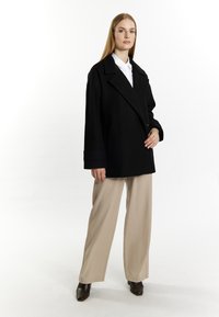 Zwarte oversized jas met een dubbele knoopsluiting, brede revers en omgeslagen mouwen, gecombineerd met lichtbeige broek en donkere enkellaarsjes.
